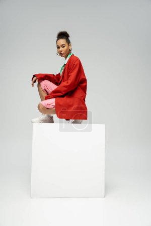 mujer afroamericana en traje de moda y vibrante sentado en la parte superior del cubo en el fondo gris