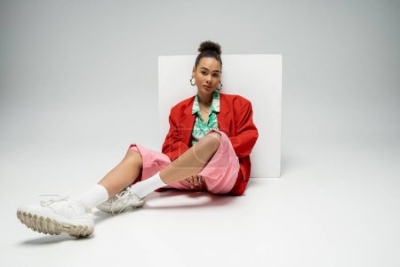pretty african american woman in fashionable and vibrant outfit sitting near cube on grey backdrop