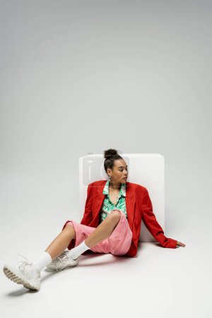 Foto de Mujer afroamericana de moda en traje de moda y vibrante sentado cerca del cubo en el fondo gris - Imagen libre de derechos