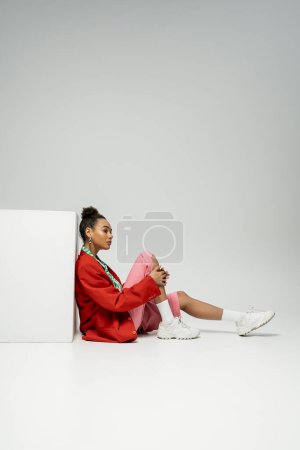 thoughtful african american woman in modern and vibrant clothes sitting near cube on grey backdrop