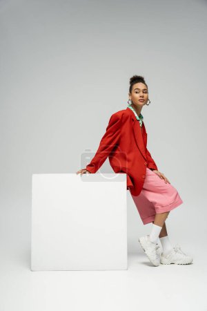 attractive african american woman in vibrant outfit leaning on cube on grey backdrop, look away