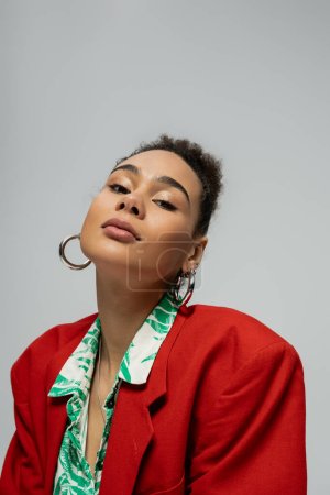 african american woman in stylish red blazer and hoop earrings looking at camera on grey backdrop