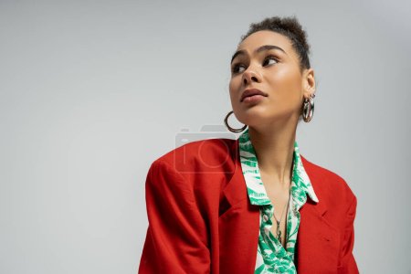 mujer afroamericana en elegante chaqueta roja y pendientes de aro mirando hacia otro lado sobre fondo gris