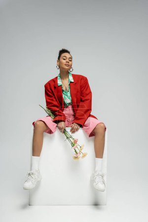 african american woman in stylish red blazer holding flowers and sitting on cube on grey backdrop