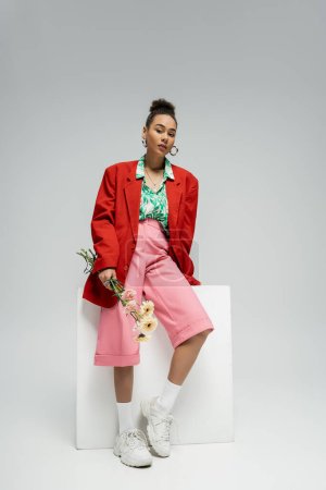 african american woman in stylish red blazer holding flowers and leaning on cube on grey backdrop