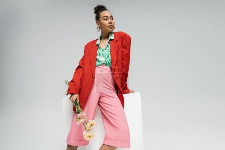 pretty african american woman in red blazer holding flowers and leaning on cube on grey backdrop