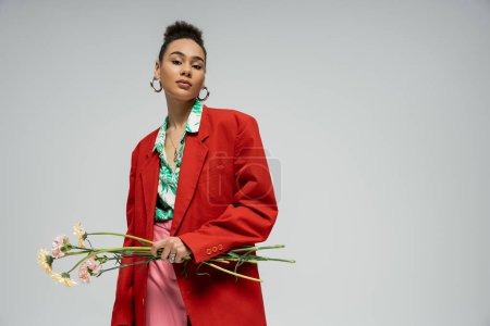 pretty african american woman in red blazer holding flowers and looking at camera on grey backdrop
