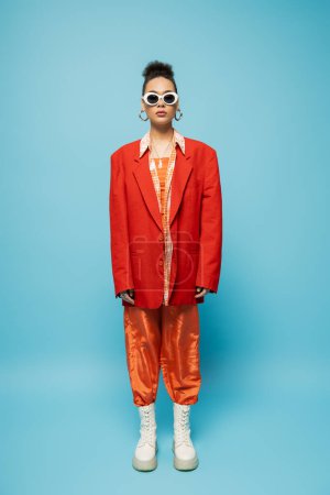 stylish african american woman in trendy sunglasses and vibrant outfit posing on blue backdrop