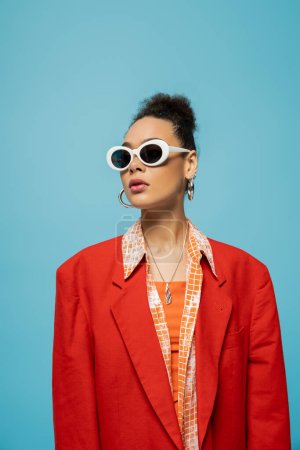 african american woman in hoop earrings, sunglasses and vibrant outfit posing on blue backdrop