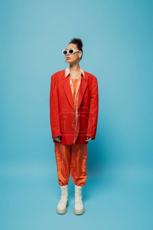 Afrikanische Amerikanerin in Sonnenbrille und pulsierendem Outfit posiert vor blauem Hintergrund