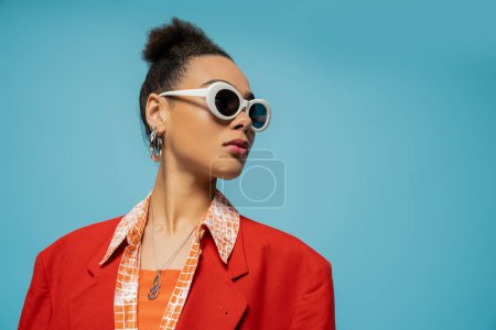 Téléchargez les photos : Portrait de femme afro-américaine en boucles d'oreilles cerceau, lunettes de soleil et tenue vibrante posant sur bleu - en image libre de droit