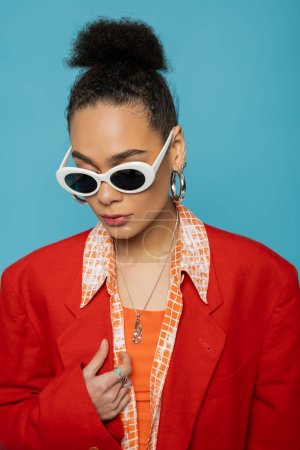 Porträt einer afrikanisch-amerikanischen Frau mit Reifrohren, Sonnenbrille und lebendigem Outfit, die auf blauem Grund posiert