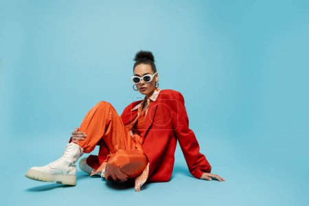 persönlicher Stil, junges afrikanisch-amerikanisches Model in lebendigem Outfit sitzt auf blauem Hintergrund