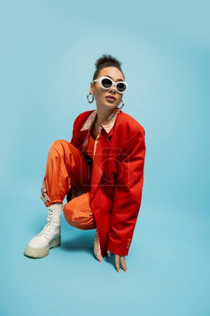 einzigartigen Stil, junge afrikanisch-amerikanische Modell in trendigen lebendigen Outfit sitzt auf blauem Hintergrund