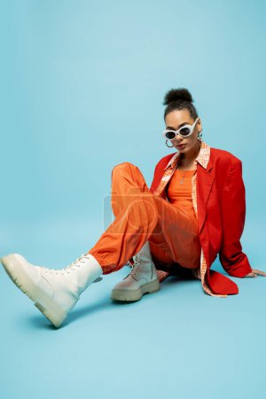 einzigartiger Stil, ausdrucksstarkes afrikanisch-amerikanisches Model im trendigen, lebendigen Outfit auf blauem Hintergrund sitzend