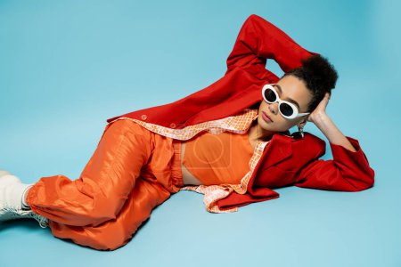 unique style, expressive african american model in trendy vibrant outfit lying on blue background