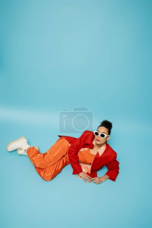 fashionista, expressive african american model in trendy vibrant outfit lying on blue background