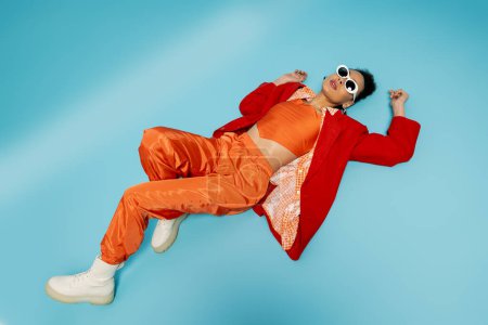 top view of trendy african american model in stylish vibrant outfit lying on blue background