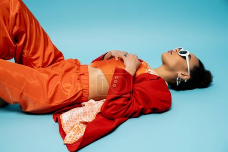 attractive african american model in stylish vibrant outfit lying on blue background, relaxed pose