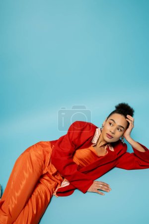Téléchargez les photos : Joli modèle afro-américain en accessoires argentés et tenue vibrante couchée sur fond bleu - en image libre de droit