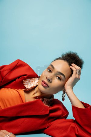 attractive african american model in silver accessories and vibrant outfit lying on blue backdrop