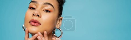 beautiful african american model in silver hoop earrings and rings looking at camera on blue, banner