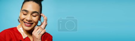 Photo for Portrait of pleased african american model in silver accessories smiling on blue backdrop, banner - Royalty Free Image