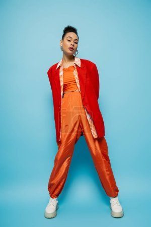 gorgeous fashion model in bright orange clothing looking at camera and posing on blue background