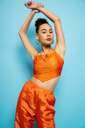 elegante mujer afroamericana en traje de calle naranja brillante posando con su mano levantada