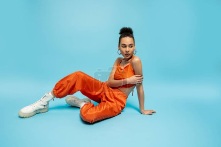 junge Afroamerikanerin in trendiger orangefarbener Kleidung mit Accessoires auf dem Boden sitzend und posierend