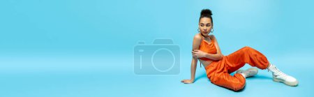 stylish african american woman in urban orange outfit with accessories sitting on floor, banner