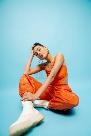 Nahaufnahme lächelndes afrikanisch-amerikanisches Model in fetter orangefarbener Kleidung, das mit der Hand auf dem Kopf auf dem Boden sitzt