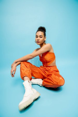 Photo for Gorgeous fashion model in vivid urban clothing sitting on floor with her arms on knee, blue backdrop - Royalty Free Image