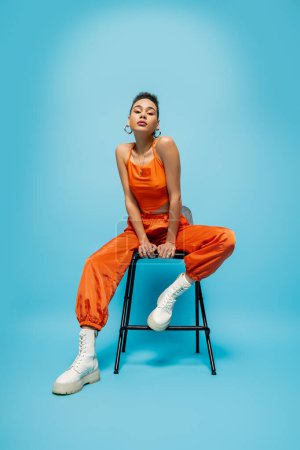 stylisches afrikanisch-amerikanisches Model sitzt auf einem Hochstuhl in lebendiger Streetwear posiert vor blauem Hintergrund