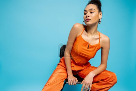 close up attractive fashion model in orange urban suit sitting on tall chair on blue backdrop