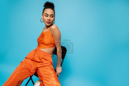 Foto de Modelo de moda hermosa en ropa naranja sentado en una silla alta mirando hacia otro lado posando sobre fondo azul - Imagen libre de derechos
