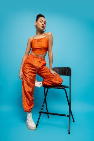 stylish african american woman in vivid clothing with earrings posing near tall chair, blue backdrop