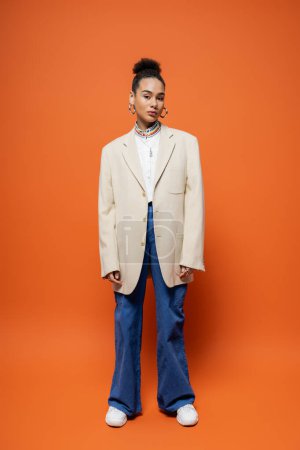stylish african american woman in urban outfit with beige blazer posing on bright orange backdrop