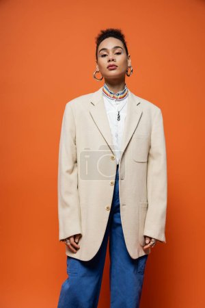Trendy junges Mode-Model in beigem Blazer und Jeans mit Reifrohrringen posiert auf orangefarbenem Hintergrund