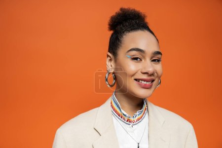 close up happy fashion model with hoop earrings looking and smiling at camera on orange background