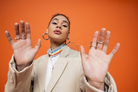 young fashion model in beige blazer with hoop earrings and silver rings on orange background