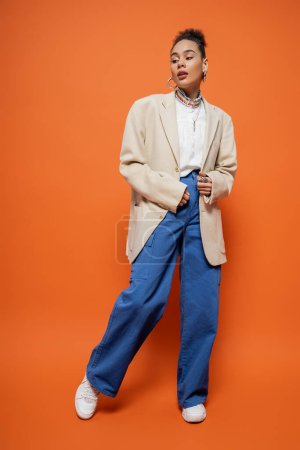 pretty african american model in stylish beige blazer and blue pants posing on orange background