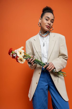 junges hübsches afrikanisch-amerikanisches Model in beigem Blazer mit Reifrohren mit Blumenstrauß