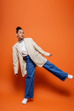 modelo de moda con clase en blazer beige y pantalones azules de pie sobre una pierna posando sobre fondo naranja
