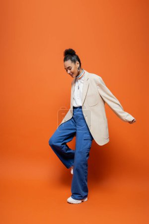 alegre modelo afroamericano de moda en chaqueta beige y pantalones azules sonriendo mirando hacia abajo