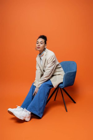 beautiful fashion model in beige blazer blue pants and bright striking makeup sitting on blue chair