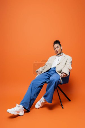 Téléchargez les photos : Beau modèle de mode afro-américain dans la rue tenue vibrante assis détendu sur chaise bleue - en image libre de droit