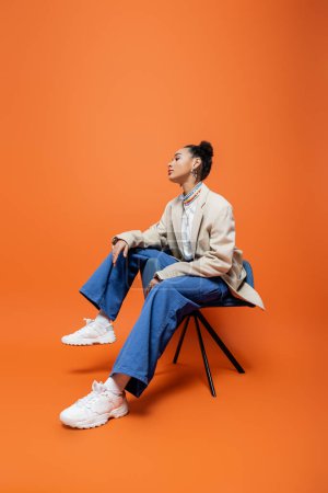 Photo for Stylish african american model in fashionable and trendy outfit sitting on blue chair looking away - Royalty Free Image