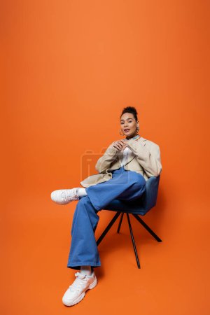 stylish african american woman in fashionable outfit sitting on blue chair with hands closer to face
