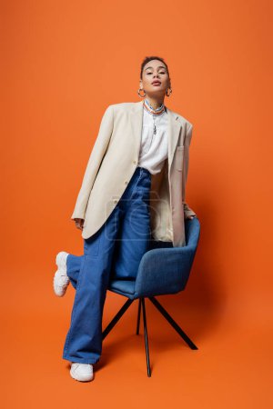 attractive african american woman in beige blazer and blue pants with vivid makeup, knee on chair